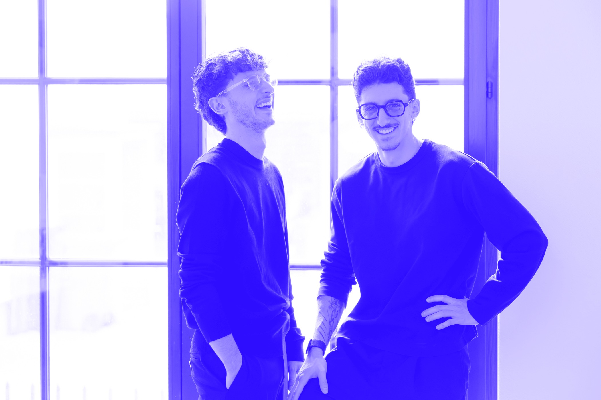 Noah und Fabiano lachen sympathisch in die Kamera vor einem Fenster, dass Bild ist mit dem Impuls+ blau eingefärbt